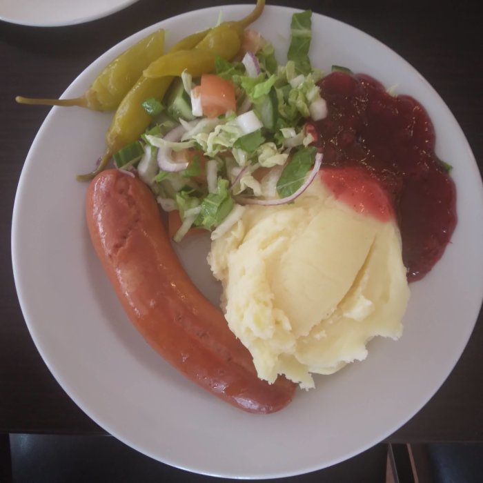 En tallrik med falukorv, potatismos, lingonsylt och sallad med blandade grönsaker och inlagda feferoni.