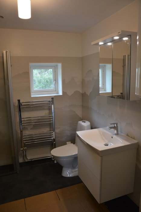 Nästan färdigt badrum med bergsmönstrad tapet, handfat, spegelskåp, toalett och handdukstork.