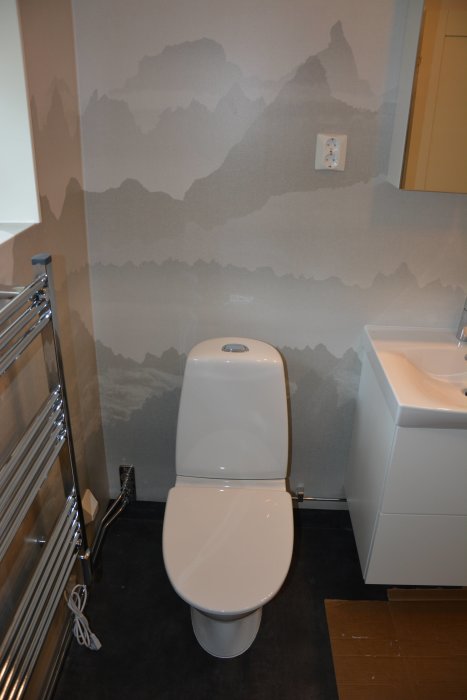 Nyrenoverat badrum med toalettstol, handduksstång och tvättställ mot vägg med bergsmönstrad tapet.