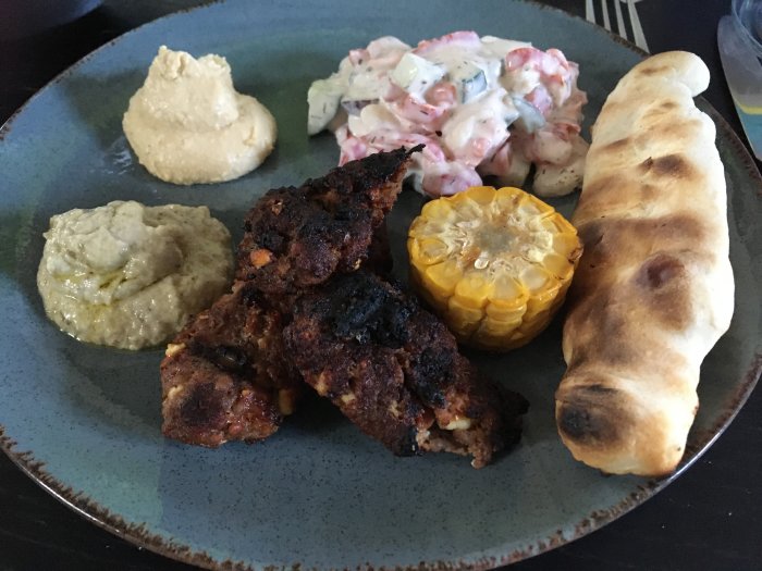 En tallrik med grillad mat: köttfärsbiffar, majs, pinnbröd, hummus, baba ganoush och potatissallad.