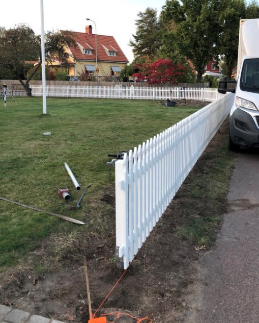 Nytt vitt staket längs en gräsmatta med hus och röda buskar i bakgrunden.