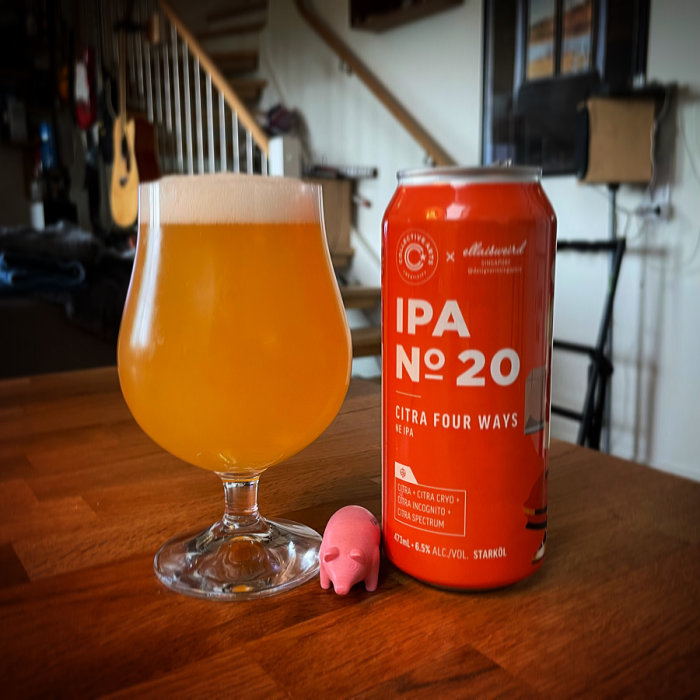 En öl i glas bredvid en burk IPA nr 20 och en liten rosa plastfigur på ett träbord.