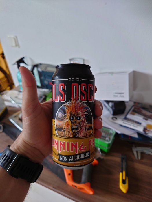 Hand håller en burk alkoholfri dryck med etikett med eldig karaktär, verktyg syns suddigt i bakgrunden.