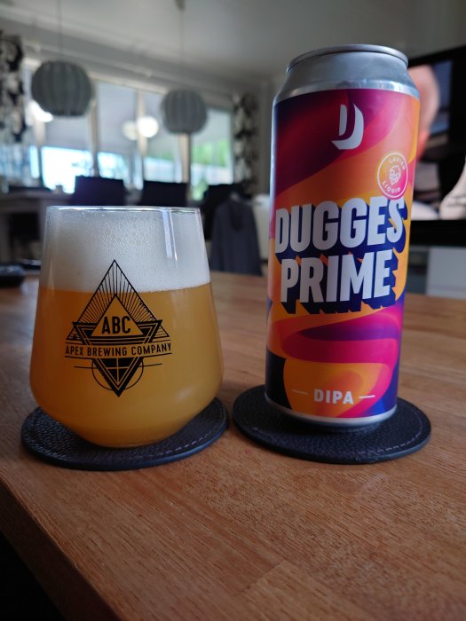 Ölglas fyllt med öl och en burk Dugges Prime DIPA på ett bord, interiör i bakgrunden.