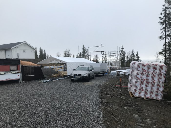 Byggplats med uppsatt tält, materialleverans, fordon och byggnadsställning i bakgrunden.