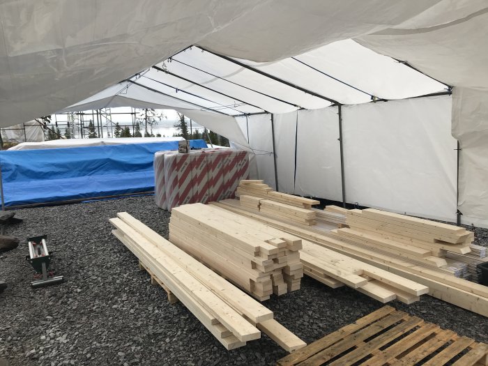 Tält på byggplats med staplade träbjälkar och verktyg, byggställning synlig i bakgrunden.