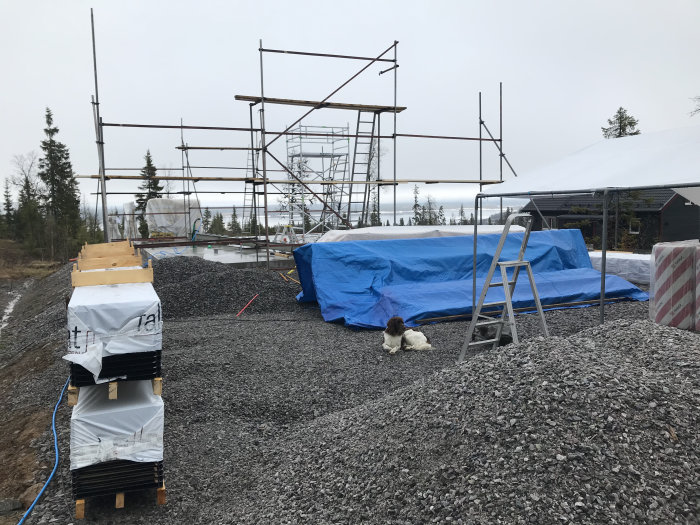 Byggplats med uppsatt tält, materialleverans och delvis rest ställning under grå himmel.