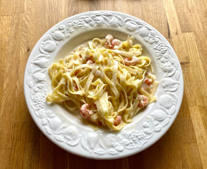 Tallrik med pasta och kräftstjärtar i vitvinssås på träunderlag.