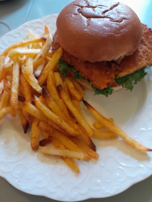 Hemgjord kycklingburgare med panerad filé, aioli, sallad, rödlök, majonnäs och rostad lök, serverad med kryddiga pommes frites.