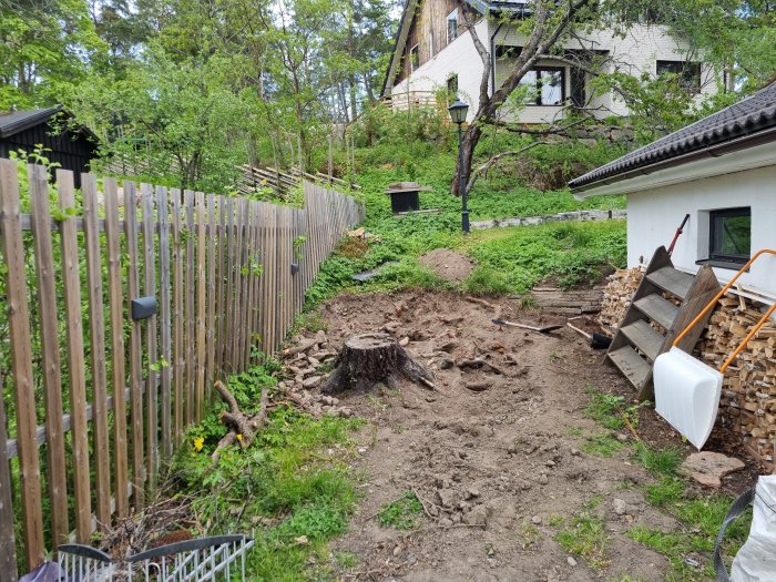 Trädgård i utgrävningsskedet med jordhögar, en stubbe, och byggmaterial vid ett hus och ett staket.