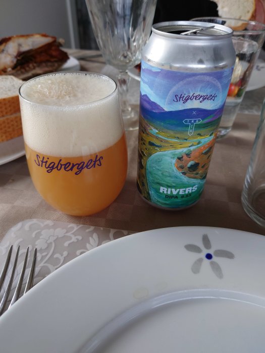 Ölglas med skum på bord bredvid ölburk med färgglad etikett, tallrik och vattenglas.