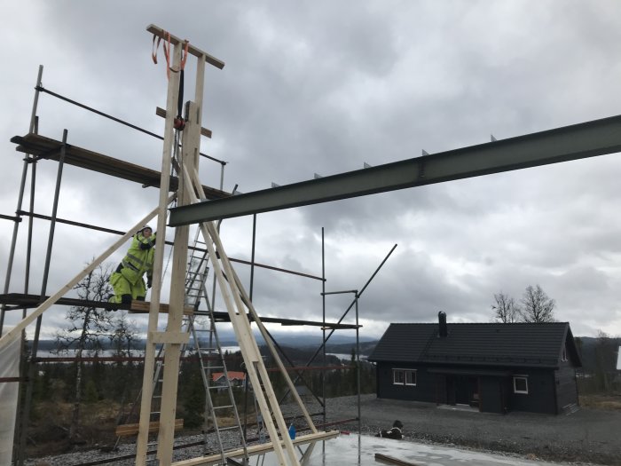 Byggplats med arbetare som monterar en nockbalk på pelare, träställningar och halvfärdig takkonstruktion, molnig himmel.