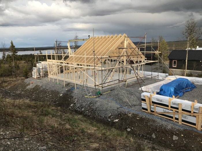 Halvfärdig trästomme av hus med resade väggar och delvis monterade takåsar, stående på pelare, omgiven av byggmaterial.