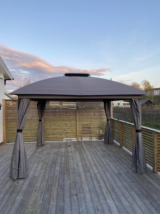 En paviljong med grått tak och gardiner på en träaltan mot en bakgrund av en solnedgångshimmel.