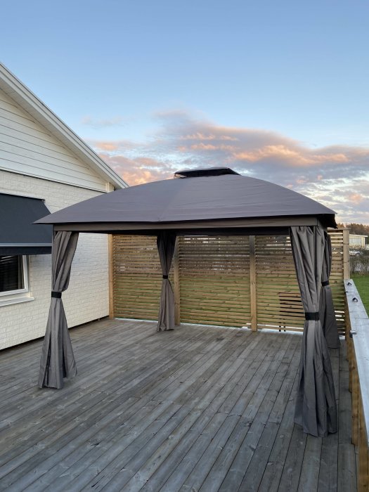 Paviljong med tak och draperier på trädäck intill vit husvägg, solnedgång i bakgrunden.