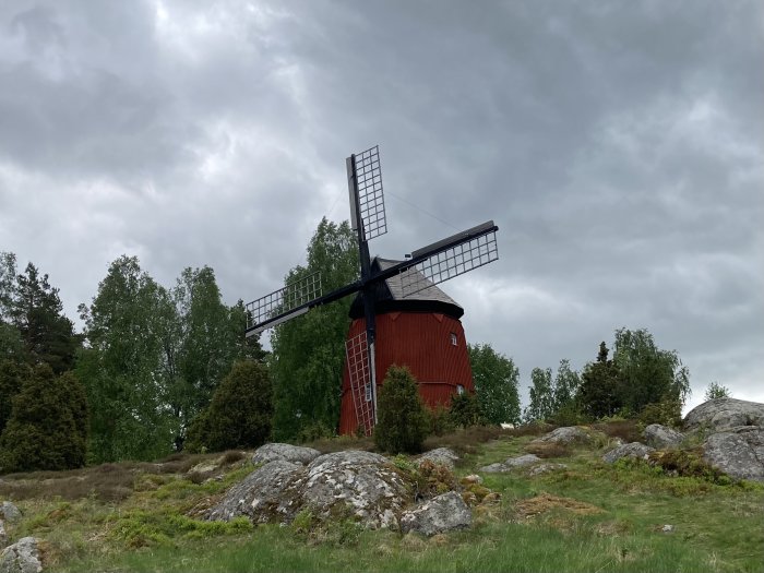 Traditionell röd väderkvarn med svarta vingar mot en molnig himmel, omgiven av grönska och stenar.