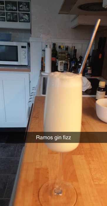 Ett glas Ramos gin fizz med tunt skum på köksbänk, bakgrund av spritflaskor.