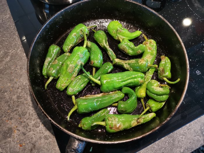 Gröna pimientos de Padron stekta i olja med salt i en svart stekpanna på spishäll.