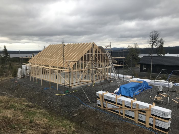 Regelstommen till ett hus under konstruktion med takåsar på plats och delvis pålagd takduk, omgiven av byggmaterial.