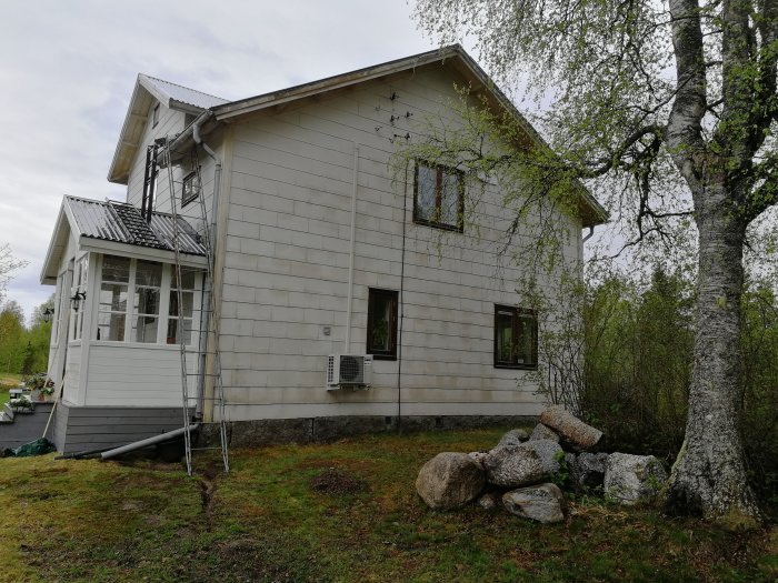 Gammalt timmerhus med eternitplattor på fasaden, stegar lutade mot huset, träd och stenar i förgrunden.