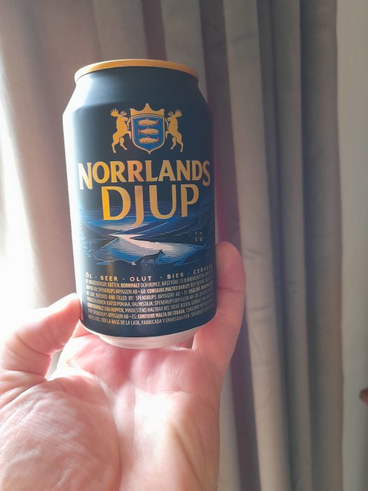 Hand håller en burk Norrlands Djup öl framför ett fönster med draperier.