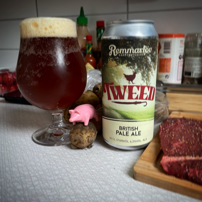 En ölglas fyllt med rödbrun öl nära en burk av Tweed British Pale Ale, med en liten rosa plastgris på en potatis.