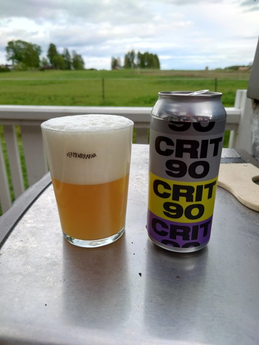 Ett glas öl med rikligt skum bredvid en burk märkt "CRIT 90" på ett utomhusbord med landskap i bakgrunden.