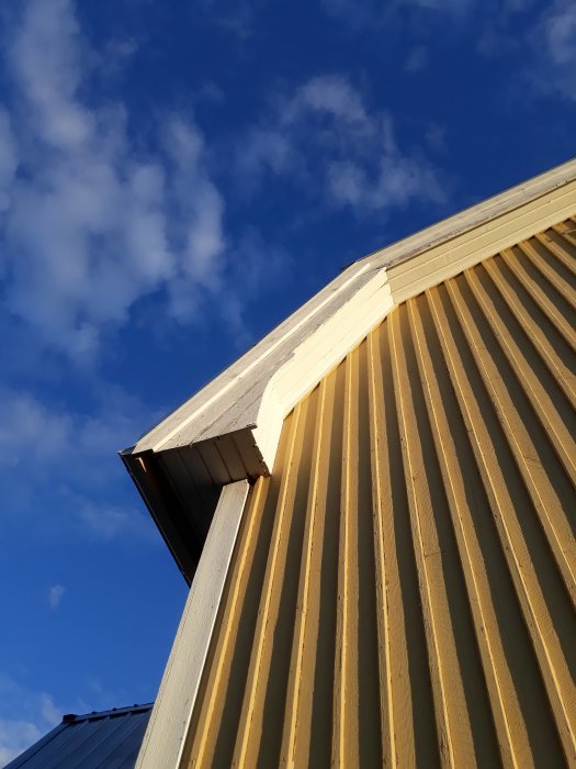 Hörnet av ett gult hus med en närbild på vindskivorna och takplåten mot en blå himmel.