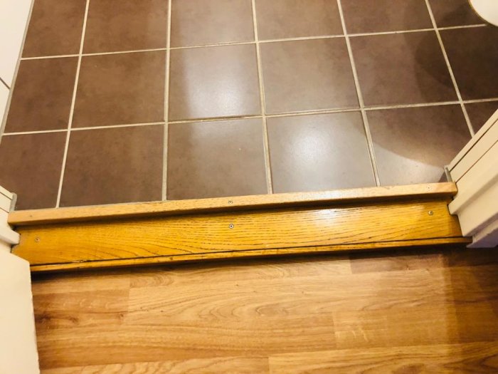 En tröskel mellan ett badrum med kakelgolv och en hall med trägolv visar en stor springa under en stängd dörr.
