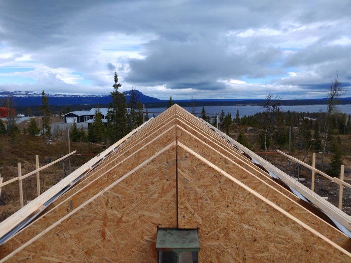 Tak under konstruktion med OSB-skivor och halvt lagt plåttak mot en bakgrund med skog och molnig himmel.