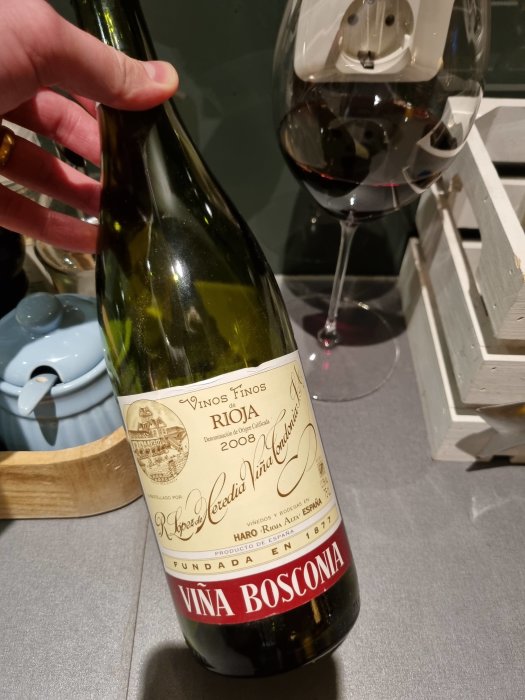 Hand håller en flaska Vina Bosconia Rioja vin från 2008, med ett glas rödvin i bakgrunden.