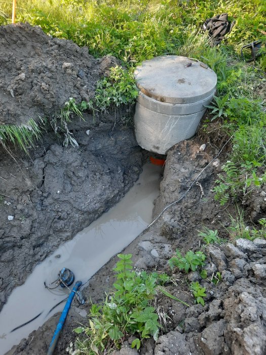 Grävd grop med vatten och avloppsrör under installation, omgiven av gräs och lera, med en svag pump.