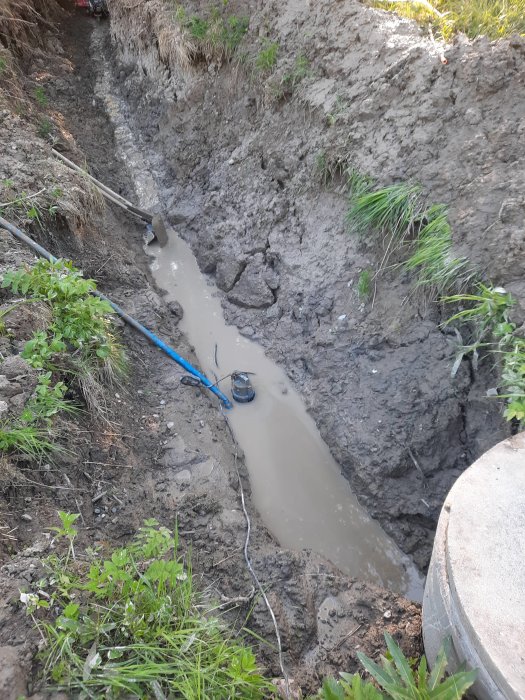 Grävd schaktgrop med vatten och en pump för att lägga avloppsrör, mark och vegetation synlig.