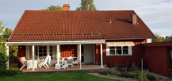 Södersida av ett hus från 1979 med rött betongtegeltak, skorsten och veranda i solljus.