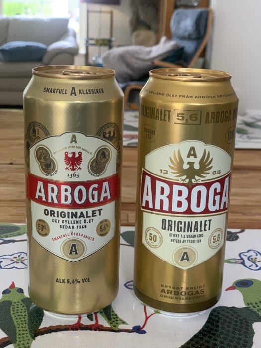 Två ölburkar från Arboga med olika designs, en med röd etikett och en med gul, står på ett bord.