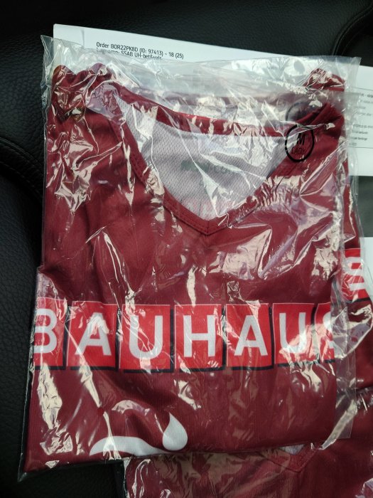 Röd t-shirt med Bauhaus-logotyp, förpackad i plast, på ett bil säte, storlek M Lady.