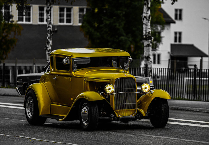 Gul hot rod-klassikerbil kör på en väg med suddig bakgrund.