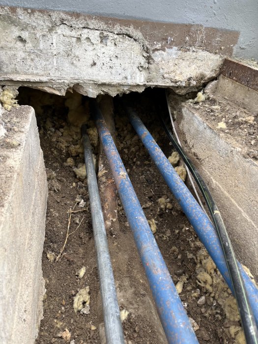 Äldre kulvert med synliga blå och grå rör samt isolering och grus, under husgrund och plåttunnel.