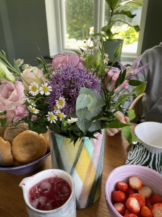 Bukett med blommor i vas på bord, omgiven av en skål med scones, jordgubbar och lemonad.