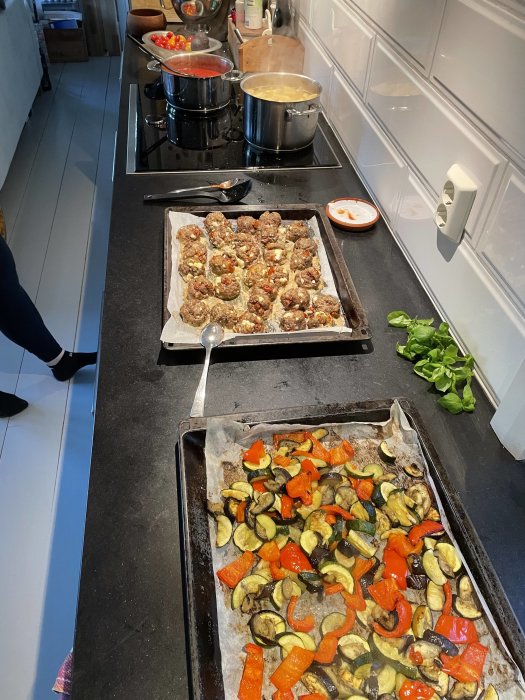 Ett kök med en bricka med köttbullar och en med rostade grönsaker, kokande pasta och sås.
