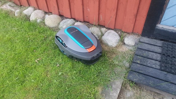 Robotgräsklippare stannad vid rödmålad stuga och stenar, indikerar felmeddelande.