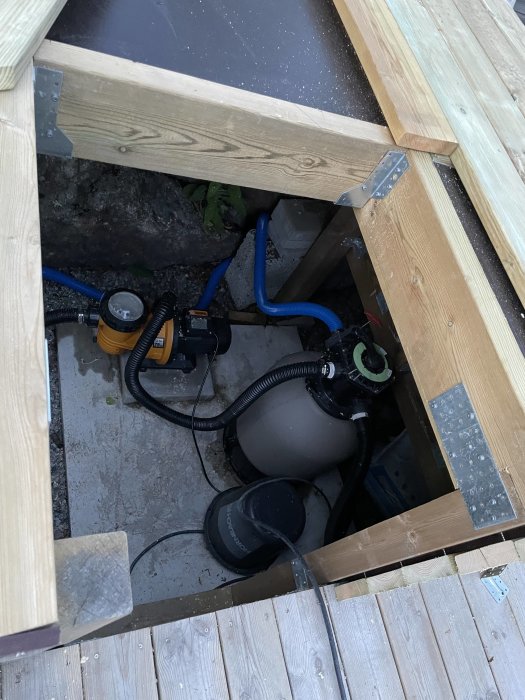 Vy under trallgolv med pump och filter, skyddad av plywood, omgiven av träram och silikonade fogar.