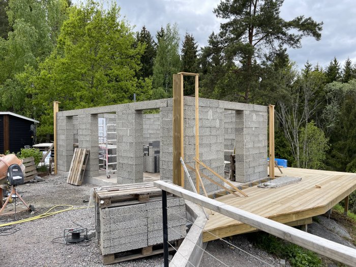 En pågående husbyggnad med gråa betongblock och nybyggt trädäck, byggmaterial och verktyg synliga.