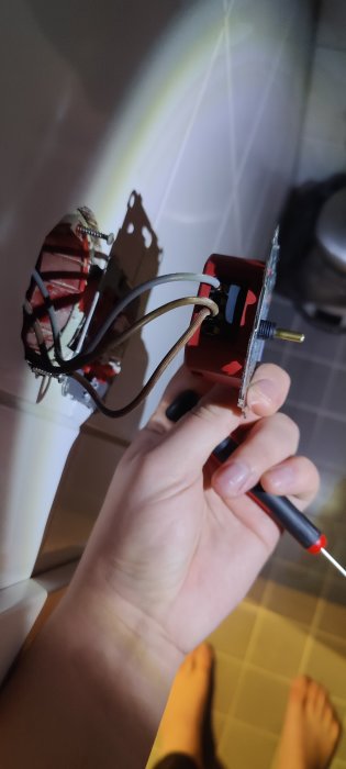 En hand håller en belysningsdimmer med exponerade kablar och skruvmejsel, i ett badrum.