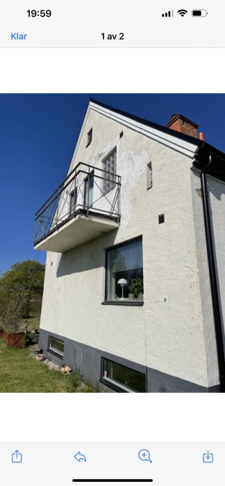 Hörnet av ett äldre hus med balkong, som visar tecken på slitage på vit putsfasad under en klarblå himmel.