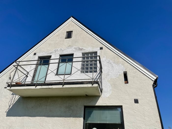 Gammalt hus med delvis skadad puts på fasaden under en balkong, behov av renovering och omarbetning.