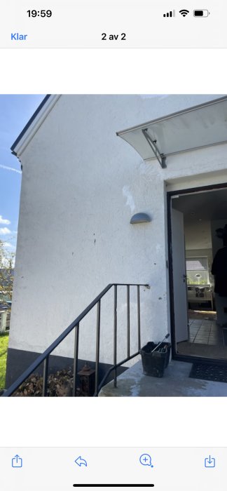 Vitputsad husfasad med synliga skador och en öppen dörr, renoveringsmaterial på trappan.