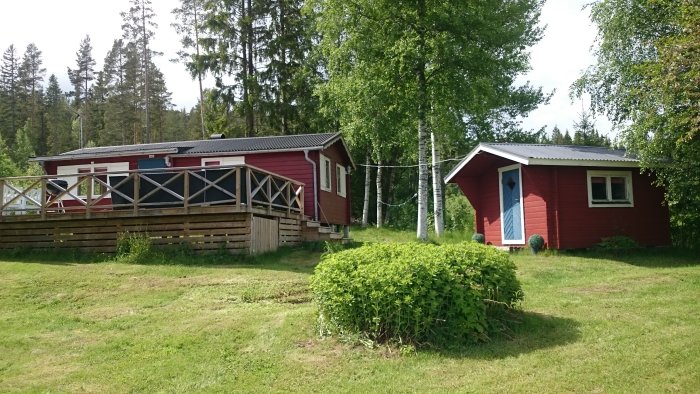 Röd stuga med vit veranda och separat uthus omgivet av grönska på plintar.