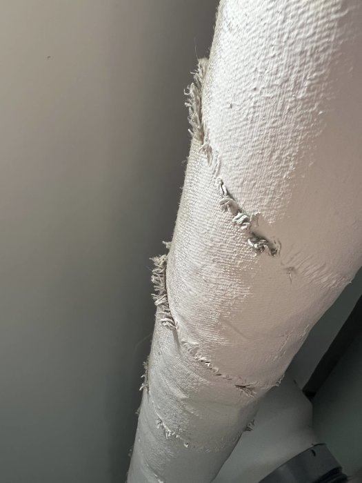 Närbild på ett slitet isolerat rör med fransiga kanter som kan vara asbest, hängande i ett tak.