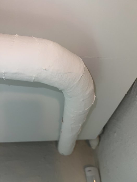 Vitmålat uppskuret rör mot en betongvägg och grått golv med synlig skarv.